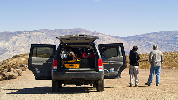 A suitcase filled suv stopped on a rural road with doors and back gate open. Two men standing outside of the car admiring the airid mountains in the distance.
